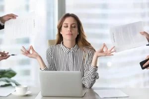 A lady is sat at her office desk with her eyes shut and hands out, relaxing. Meanwhile she's dealing with the stress of several hands in the edge of the image that are trying to pass her work to do.